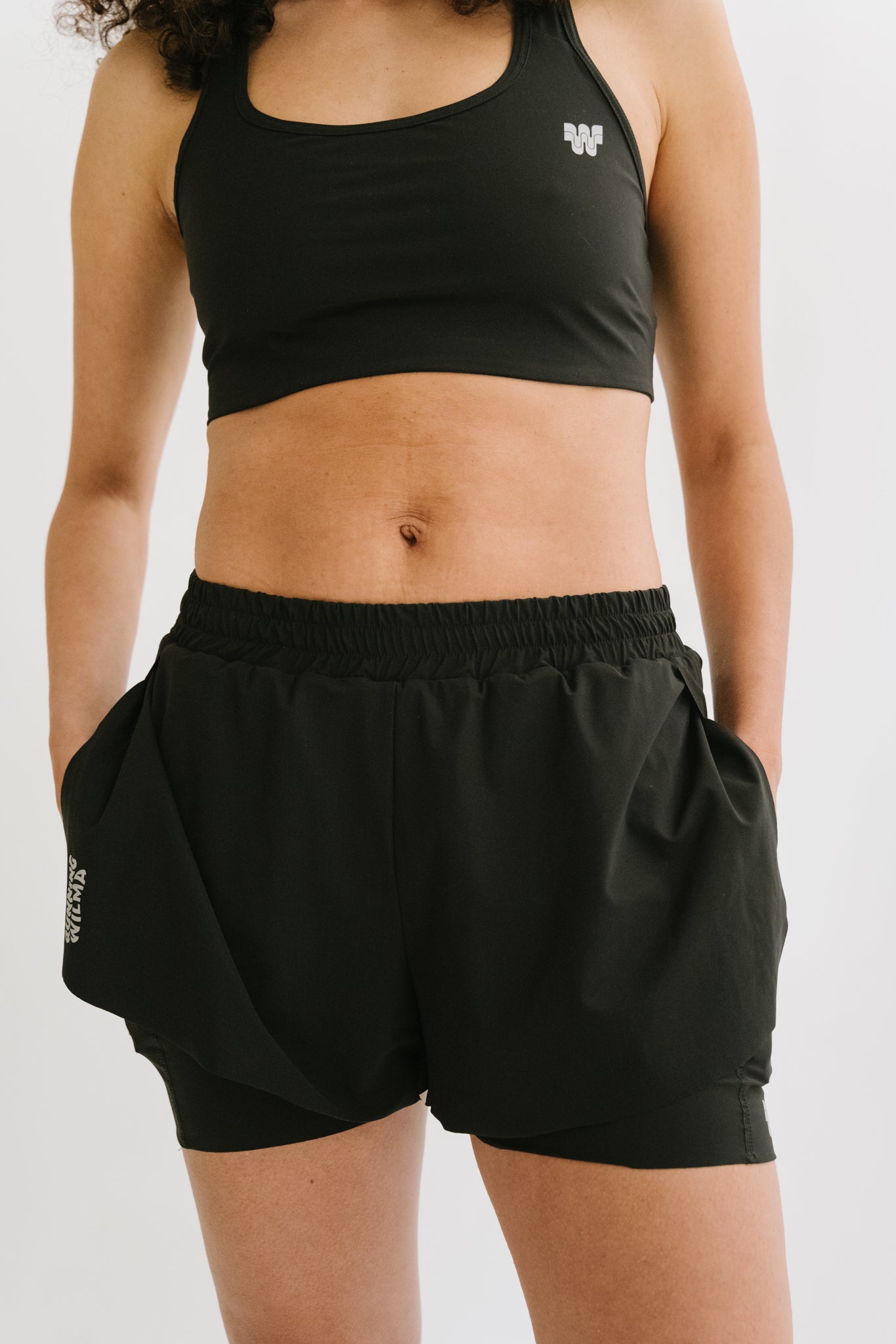 Une femme démontrant l'utilité et l'accessibilité des poches du short de running Rachel en mettant ses mains dans les deux poches latérales. #color_Noir