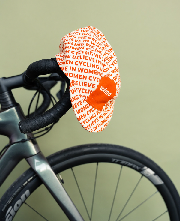 Casquette de cyclisme blanche et orange avec écrit We Believe In Women Cycling posée sur un vélo